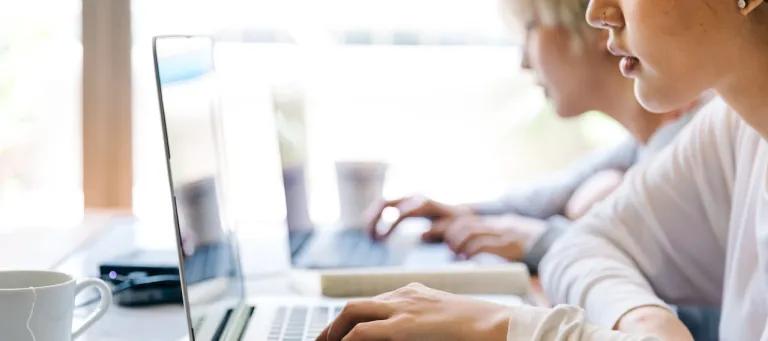 Side on photo of a woman looking at and typing on a laptop with a mug behind the laptop. 